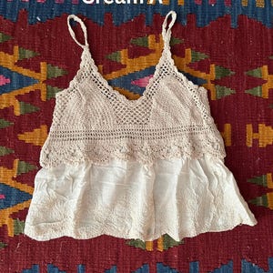 May include: Cream-coloured crochet top with spaghetti straps and a V-neck design. The top features a crocheted bodice and a flowing, embroidered bottom. The text "Cream A" is visible in the image. The top is likely a bohemian or vintage style.