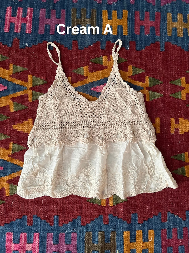May include: Cream-coloured crochet top with spaghetti straps and a V-neck design. The top features a crocheted bodice and a flowing, embroidered bottom. The text "Cream A" is visible in the image. The top is likely a bohemian or vintage style.