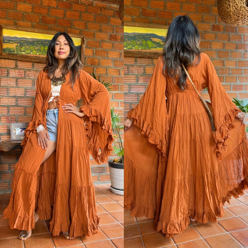 Op de afbeelding: Een oranje maxi-jurk met lange, uitlopende mouwen en ruches. De jurk is aan de voorkant open en onthult een witte top en een spijkerbroek. De jurk heeft een vloeiende, bohemien stijl.