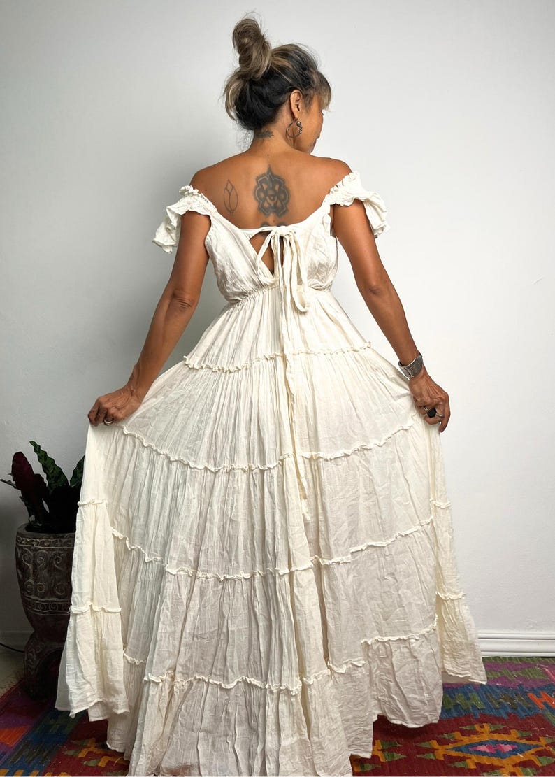 May include: Off-white tiered maxi dress with an off-the-shoulder neckline and ruffled sleeves. The dress features a tie detail at the back and a flowing, layered skirt. The dress is made of a lightweight fabric, perfect for a bohemian or summer look.