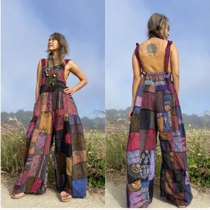 Op de afbeelding: Een patchwork jumpsuit in tinten rood, paars, bruin en blauw. De jumpsuit met wijde pijpen heeft een verscheidenheid aan vierkanten van bedrukte stof die aan elkaar zijn genaaid. De top is mosterdgeel met bordeauxrode bandjes. Het model staat in een veld.