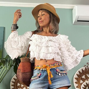 Pode incluir: Top branco com ombros descobertos com babados em camadas e mangas sino. A modelo usa shorts jeans, um chap&eacute;u de palha e um cinto colorido. Uma bolsa de tecido &eacute; usada no ombro.