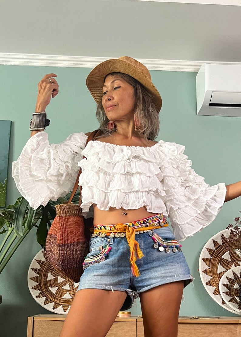 Pode incluir: Top branco com ombros descobertos com babados em camadas e mangas sino. A modelo usa shorts jeans, um chap&eacute;u de palha e um cinto colorido. Uma bolsa de tecido &eacute; usada no ombro.