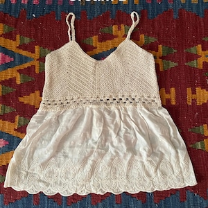 May include: Cream-coloured camisole top with a V-neck and spaghetti straps. The top features a crocheted bodice and a flowing, embroidered bottom hem. The text "Cream B" is visible in the image. The top is likely made of cotton or a similar lightweight fabric.
