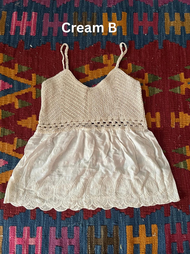 May include: Cream-coloured camisole top with a V-neck and spaghetti straps. The top features a crocheted bodice and a flowing, embroidered bottom hem. The text "Cream B" is visible in the image. The top is likely made of cotton or a similar lightweight fabric.