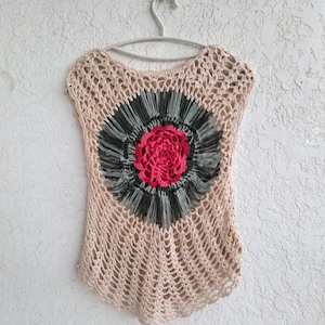May include: A crocheted top with a large, colorful flower design in the center. The top is beige with a green and pink flower.