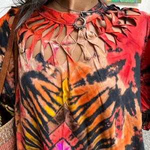 May include: A red, orange, and black tie-dye shirt with a cut-out design around the neckline. The shirt has a silver necklace with a circular pendant.
