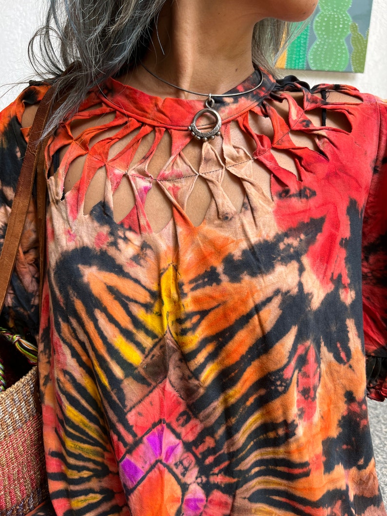 May include: A red, orange, and black tie-dye shirt with a cut-out design around the neckline. The shirt has a silver necklace with a circular pendant.
