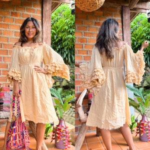 May include: A woman wearing a light beige, flowy dress with ruffled sleeves. The dress has a cinched waist and a slightly flared skirt. The woman is standing in front of a brick wall with greenery in the background.