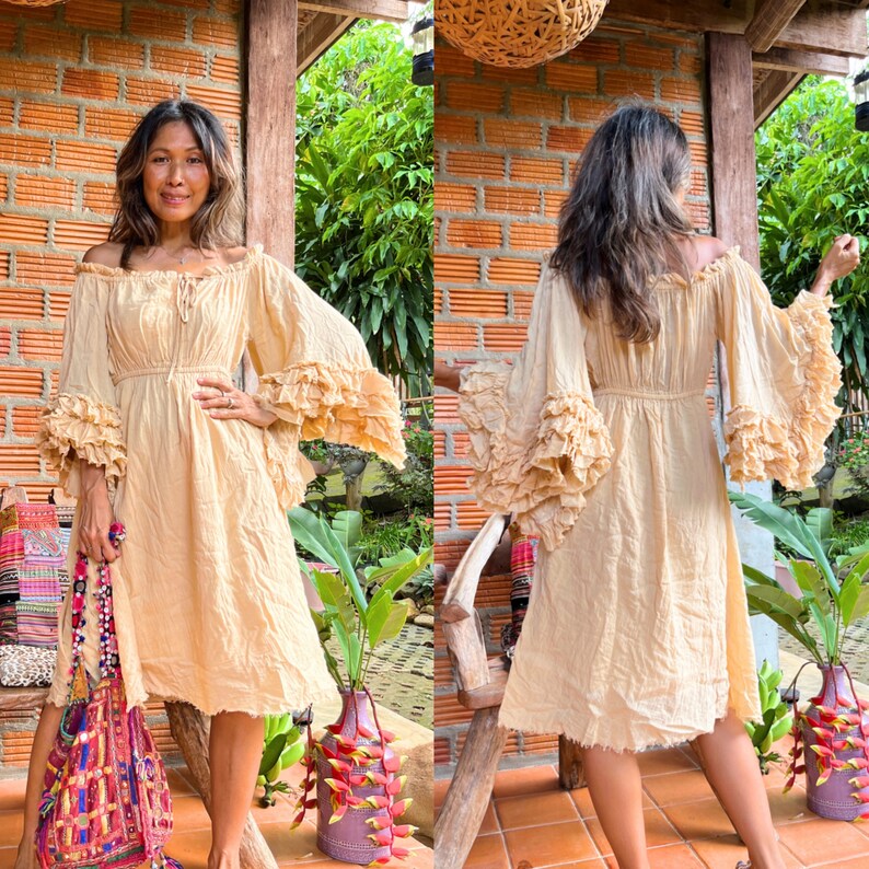May include: A woman wearing a light beige, flowy dress with ruffled sleeves. The dress has a cinched waist and a slightly flared skirt. The woman is standing in front of a brick wall with greenery in the background.