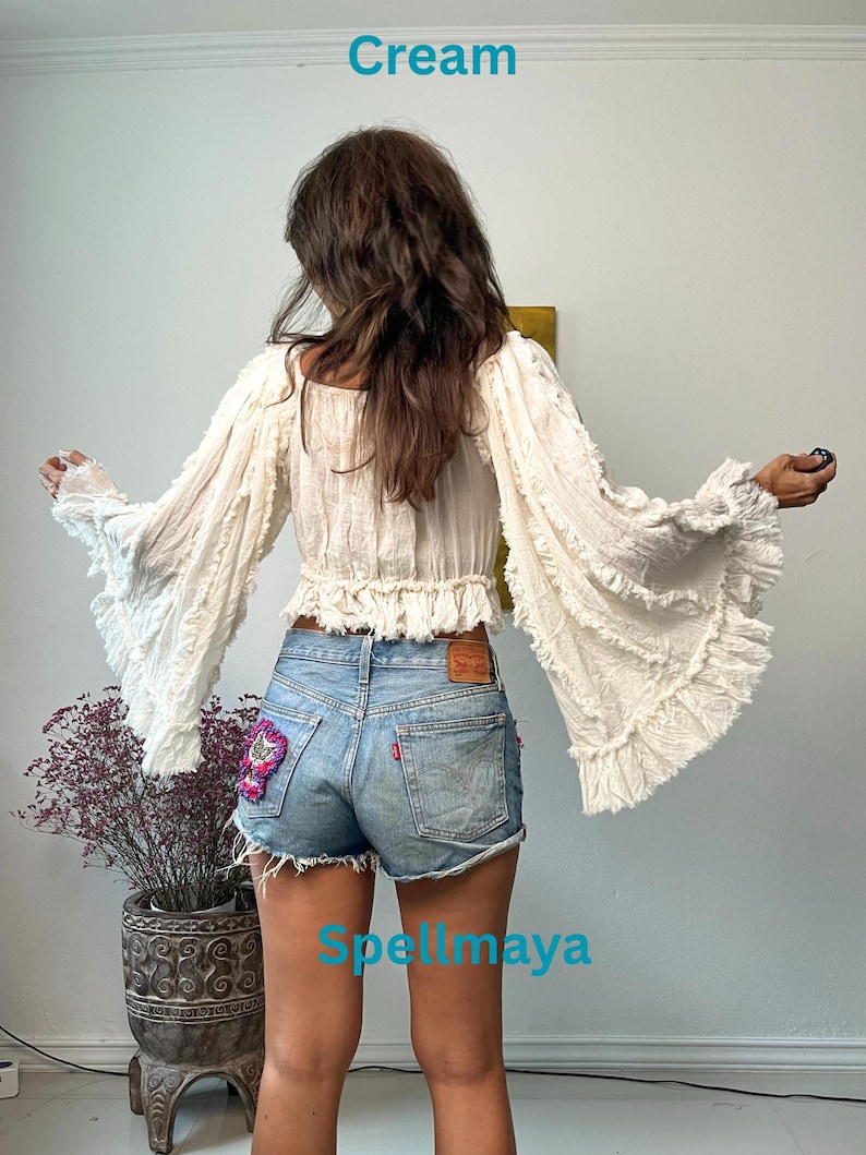 K&ouml;nnte beinhalten: Cremefarbenes Oberteil mit langen &Auml;rmeln und ger&uuml;schten Glocken&auml;rmeln. Das Oberteil wird mit Jeansshorts kombiniert. Das Wort "Cream" steht oben links. Der Text "Spellmaya" steht unten in der Mitte.