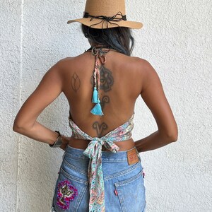 K&ouml;nnte beinhalten: Eine Frau tr&auml;gt ein hellblaues und rosafarbenes Paisley-Muster-Halter-Top mit t&uuml;rkisfarbenen Quasten, die hinten gebunden sind. Sie tr&auml;gt Jeans-Shorts mit einer Perlenverzierung auf der Ges&auml;&szlig;tasche und einen braunen Strohhut.