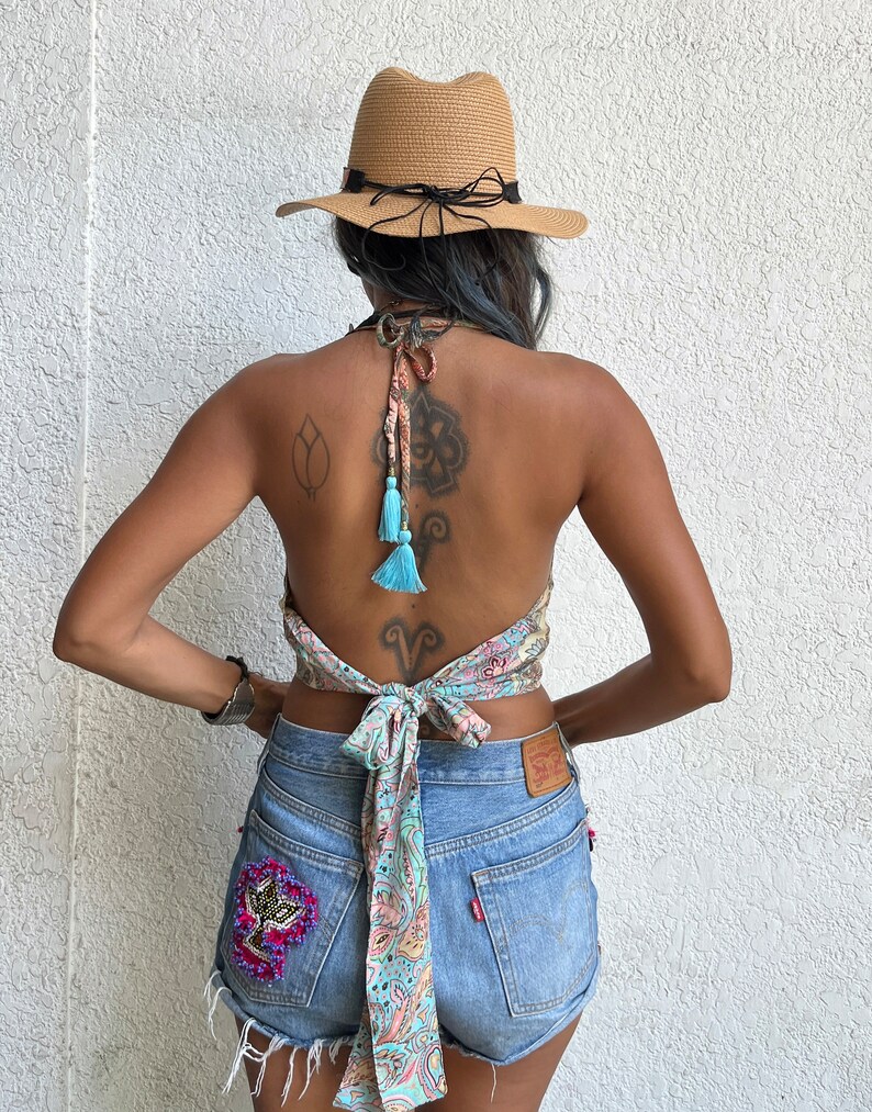 K&ouml;nnte beinhalten: Eine Frau tr&auml;gt ein hellblaues und rosafarbenes Paisley-Muster-Halter-Top mit t&uuml;rkisfarbenen Quasten, die hinten gebunden sind. Sie tr&auml;gt Jeans-Shorts mit einer Perlenverzierung auf der Ges&auml;&szlig;tasche und einen braunen Strohhut.