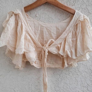 May include: A cream-colored, ruffled crop top with a tie at the front. The top is made of a lightweight fabric and has a relaxed fit.