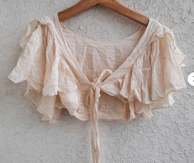 May include: A cream-colored, ruffled crop top with a tie at the front. The top is made of a lightweight fabric and has a relaxed fit.