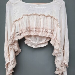 May include: A cream-colored, long-sleeved, crochet top with a ruffled neckline and fringe detailing. The top has a fitted bodice and a loose, flowing silhouette. The text "Spellmaya" is printed on the front of the top.