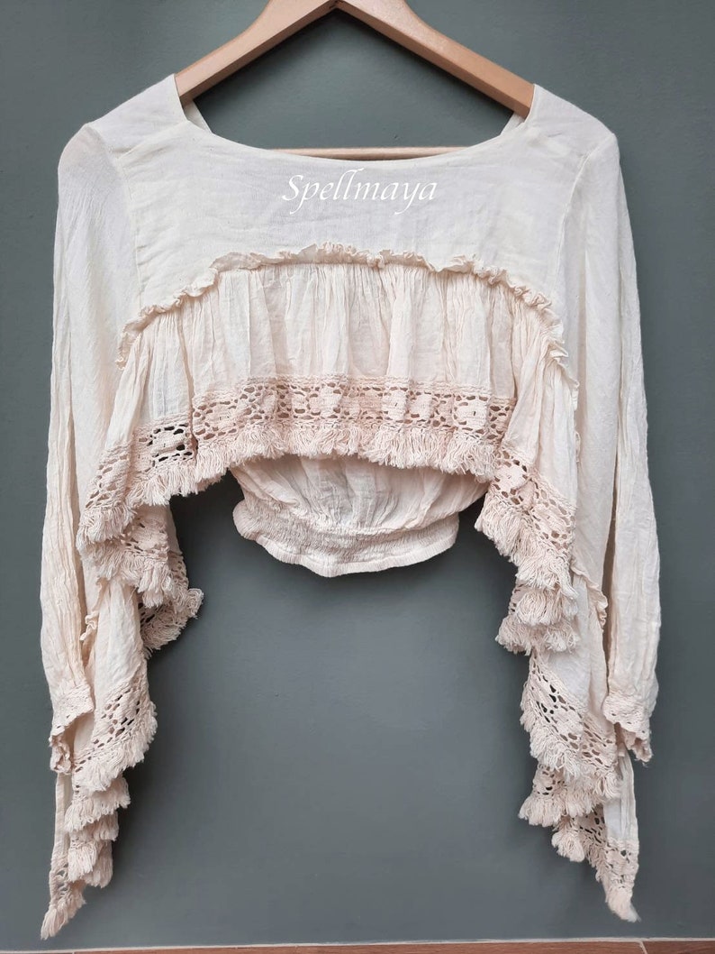 May include: A cream-colored, long-sleeved, crochet top with a ruffled neckline and fringe detailing. The top has a fitted bodice and a loose, flowing silhouette. The text "Spellmaya" is printed on the front of the top.