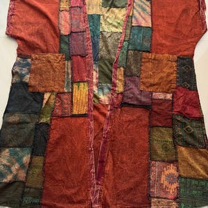 May include: A patchwork kimono-style jacket featuring the text Style BU6 at the top. The garment is constructed from various fabric squares in shades of rust, olive green, deep blue, and gold, with two large front pockets and short sleeves.