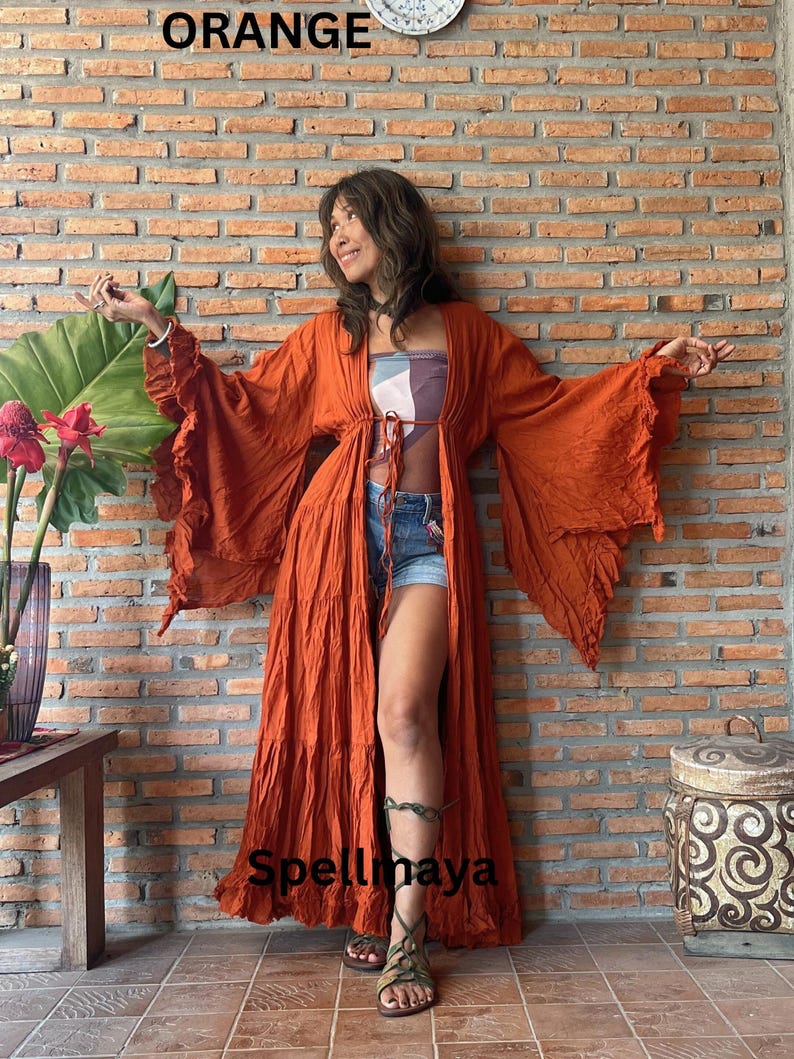 Op de afbeelding: Een oranje, lange mouwen, open voorkant robe met gerimpelde mouwen. De robe wordt gedragen over een top met patroon en een spijkerbroek. Het woord "ORANGE" staat bovenaan de afbeelding en het woord "Spellmaya" staat onderaan.