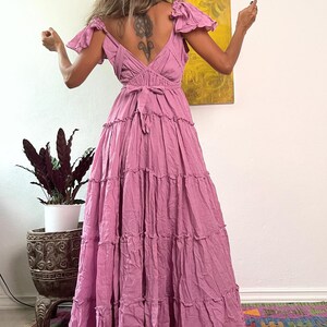 May include: A long, flowing, dusty rose dress with a tiered skirt and flutter sleeves. The dress features a V-neckline and a tie-back detail. The model is standing in front of a colorful rug and a potted plant.