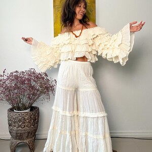 Pode incluir: Conjunto bo&ecirc;mio de duas pe&ccedil;as em branco sujo. O top &eacute; um top cropped com ombros de fora com babados em camadas e mangas compridas e largas. As cal&ccedil;as s&atilde;o de perna larga com v&aacute;rios n&iacute;veis de babados. O conjunto &eacute; feito de tecido leve.