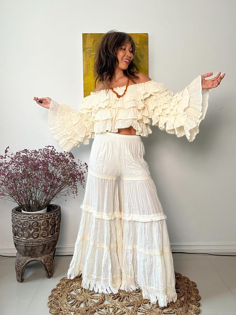 Pode incluir: Conjunto bo&ecirc;mio de duas pe&ccedil;as em branco sujo. O top &eacute; um top cropped com ombros de fora com babados em camadas e mangas compridas e largas. As cal&ccedil;as s&atilde;o de perna larga com v&aacute;rios n&iacute;veis de babados. O conjunto &eacute; feito de tecido leve.