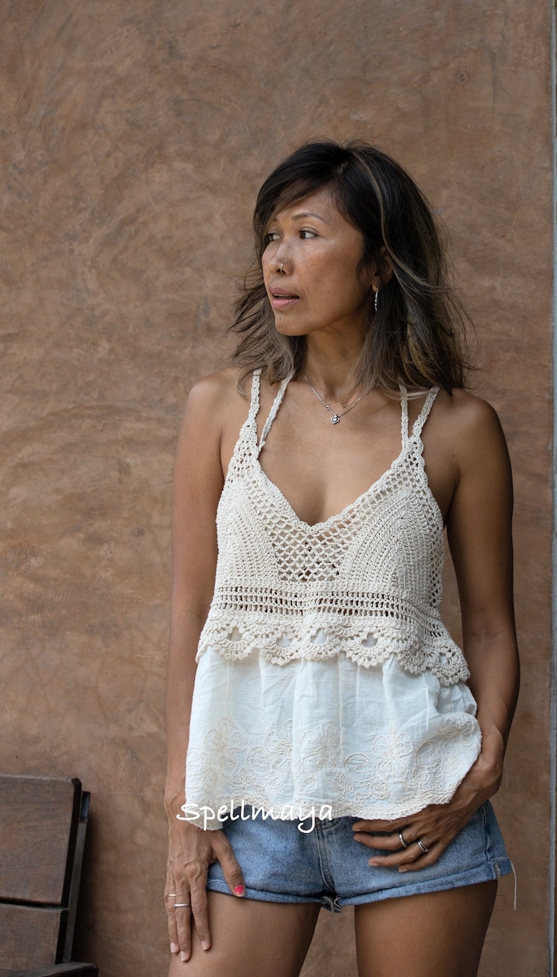 May include: A cream-coloured crochet top with a V-neck and delicate straps. The top is paired with a white embroidered blouse and denim shorts. The image is set against a brown textured wall.