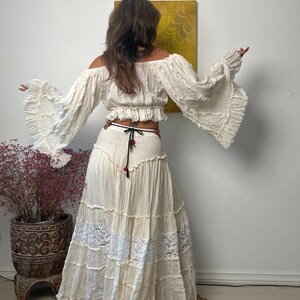 K&ouml;nnte beinhalten: Cremefarbenes Off-Shoulder-Top mit langen, ausgestellten &Auml;rmeln und einem passenden Stufenrock. Der Rock hat Spitzeneins&auml;tze und einen schwarz-roten Bindeg&uuml;rtel. Der Text "SET S" ist sichtbar.