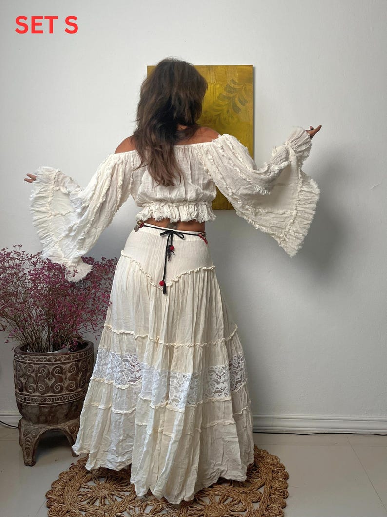 K&ouml;nnte beinhalten: Cremefarbenes Off-Shoulder-Top mit langen, ausgestellten &Auml;rmeln und einem passenden Stufenrock. Der Rock hat Spitzeneins&auml;tze und einen schwarz-roten Bindeg&uuml;rtel. Der Text "SET S" ist sichtbar.