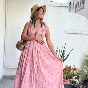 May include: A woman wearing a pink maxi dress with a V-neckline and a straw hat. She is standing on a patio with plants in the background.