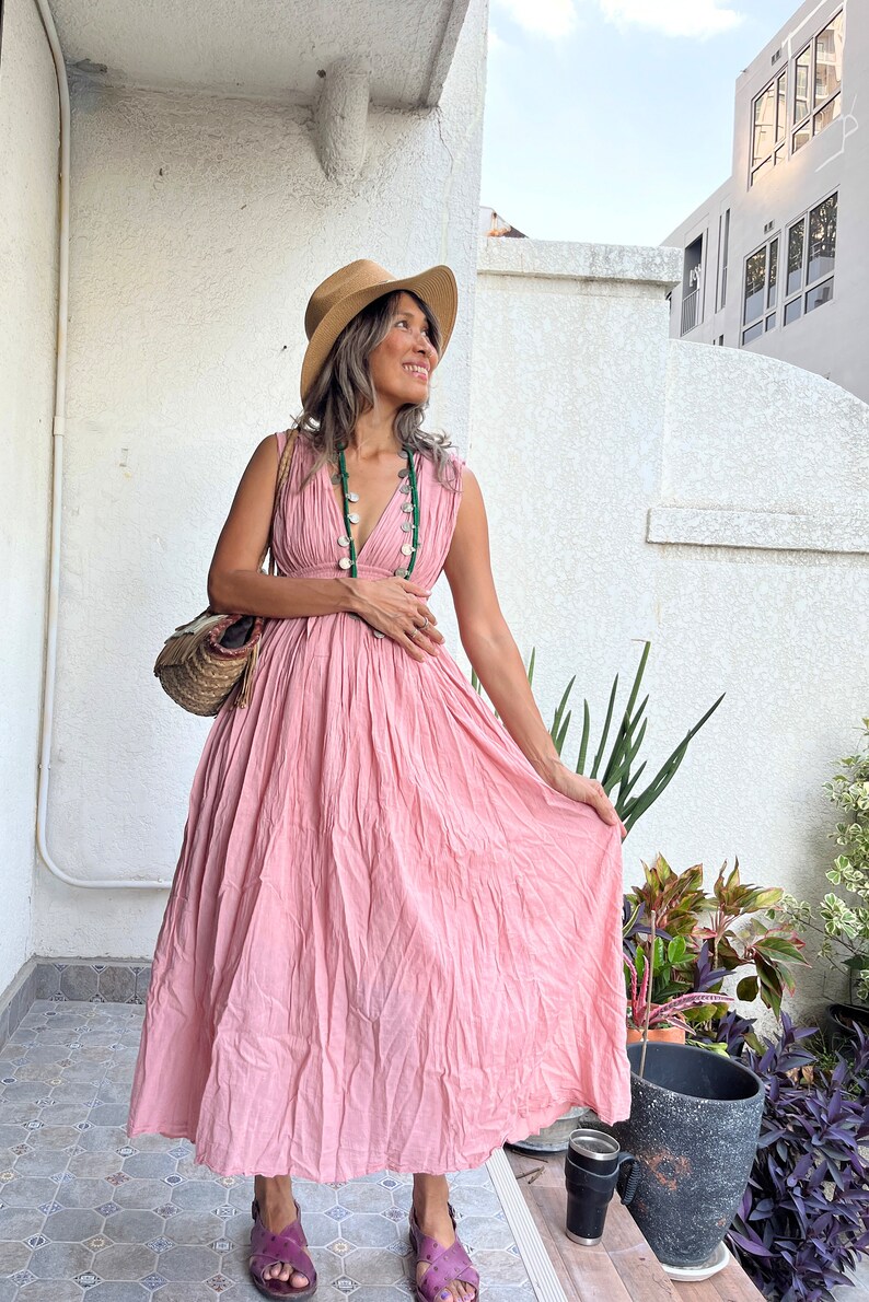 May include: A woman wearing a pink maxi dress with a V-neckline and a straw hat. She is standing on a patio with plants in the background.