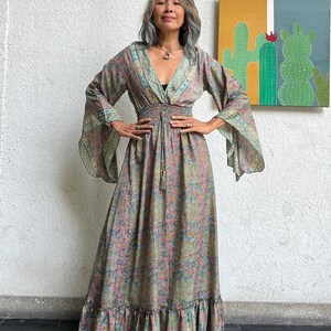 May include: A long, flowing maxi dress with a floral print in shades of pink, green, and blue. The dress has a V-neckline, long sleeves, and a cinched waist with a tie. The dress has a ruffled hem.