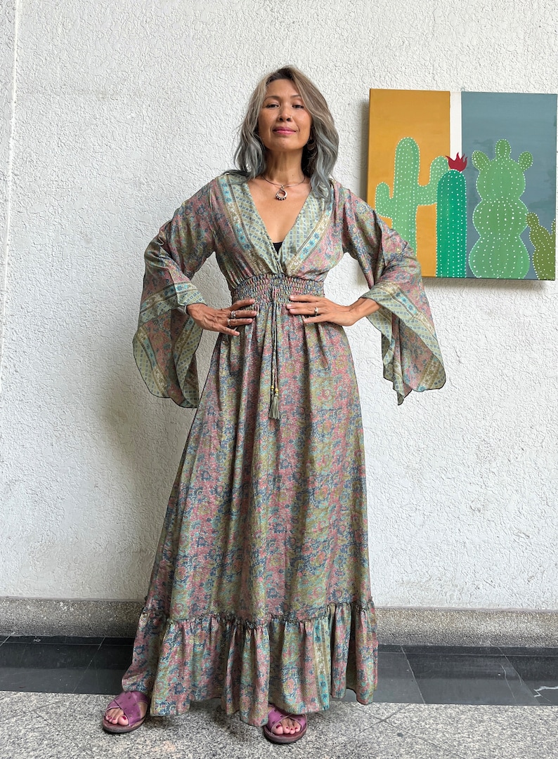 May include: A long, flowing maxi dress with a floral print in shades of pink, green, and blue. The dress has a V-neckline, long sleeves, and a cinched waist with a tie. The dress has a ruffled hem.