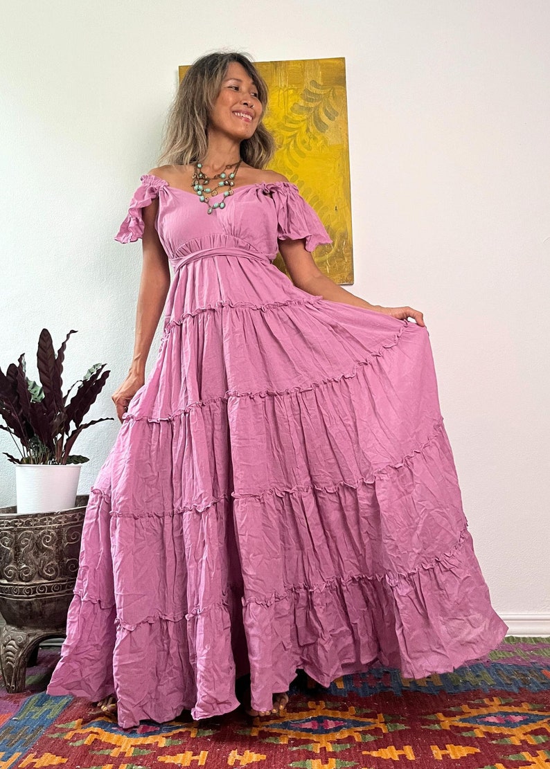 May include: A long, flowing, dusty rose-colored dress with an off-the-shoulder neckline and tiered skirt. The dress features ruffled detailing and a fitted bodice. A turquoise necklace adds a pop of color. The dress is suitable for a bohemian or casual style.