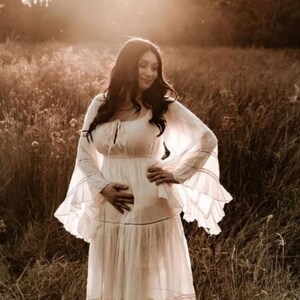May include: A pregnant woman in a white maxi dress with lace detailing and ruffled sleeves stands in a field of tall grass. The sun is shining brightly behind her, creating a warm glow.