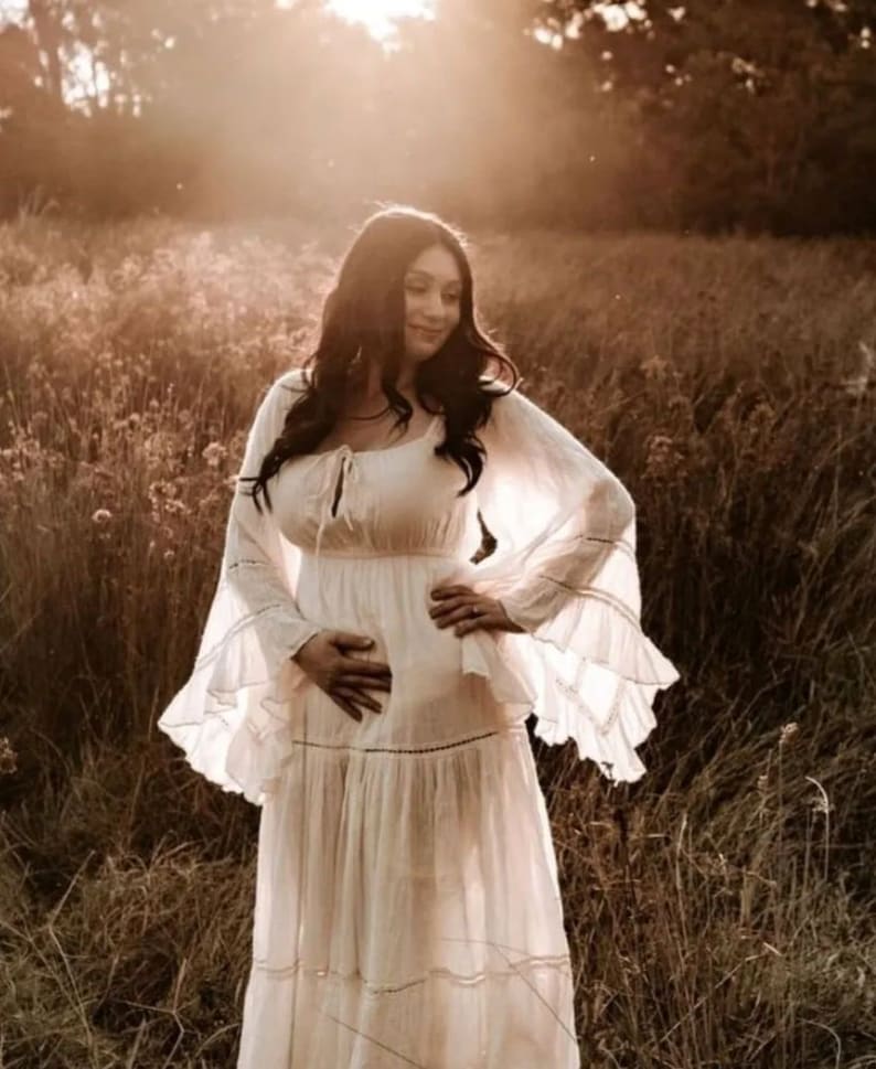 May include: A pregnant woman in a white maxi dress with lace detailing and ruffled sleeves stands in a field of tall grass. The sun is shining brightly behind her, creating a warm glow.