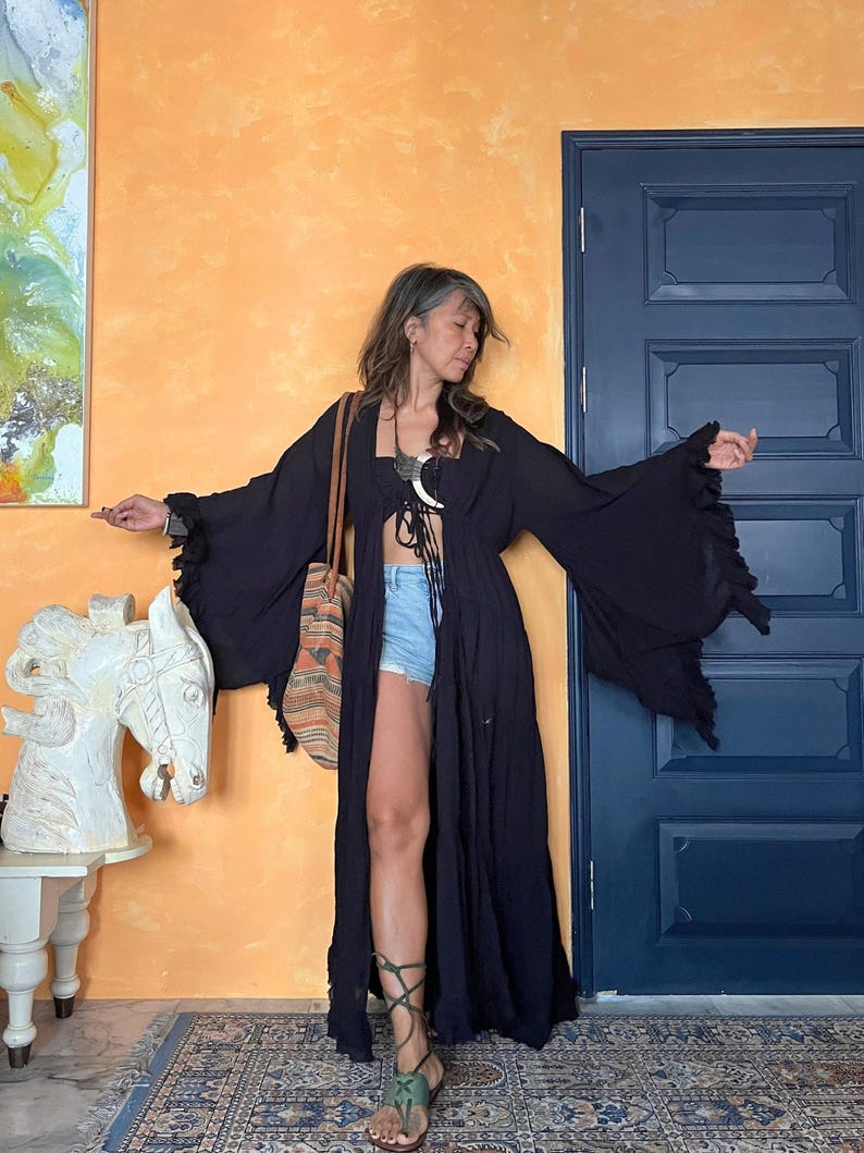 Op de afbeelding: Een zwarte kimono-achtige robe met lange mouwen en uitlopende mouwen. De robe is aan de voorkant open en wordt gedragen met een top met patroon en een spijkerbroek. Een tas met patroon wordt over de schouder gedragen. De persoon draagt groene sandalen met bandjes.