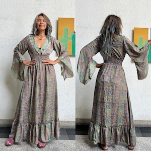 May include: A long, flowing maxi dress with a floral print in shades of pink, green, and blue. The dress has a V-neckline, long sleeves, and a gathered waist. The sleeves are bell-shaped and have a floral trim. The dress has a tiered skirt with a ruffled hem.