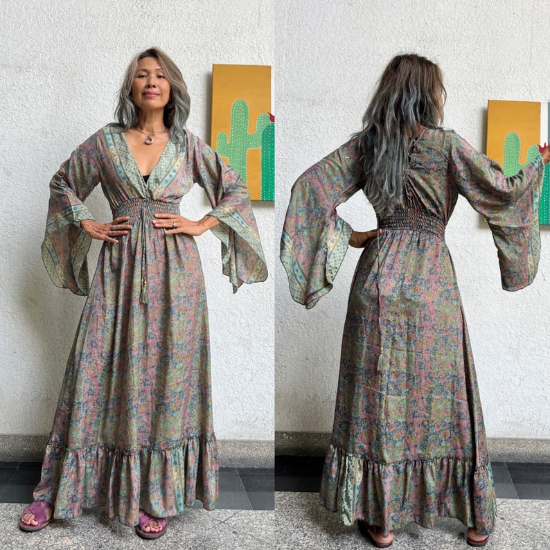 May include: A long, flowing maxi dress with a floral print in shades of pink, green, and blue. The dress has a V-neckline, long sleeves, and a gathered waist. The sleeves are bell-shaped and have a floral trim. The dress has a tiered skirt with a ruffled hem.