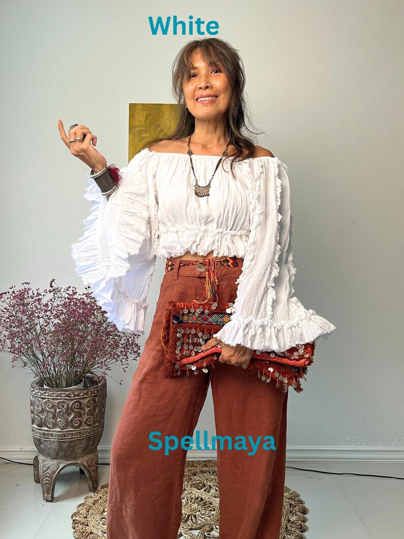 K&ouml;nnte beinhalten: Ein wei&szlig;es Off-Shoulder-Top mit R&uuml;schen&auml;rmeln und eine rostfarbene Hose mit weitem Bein. Das Outfit wird mit einer dekorativen Clutch und Schmuck erg&auml;nzt. Das Wort "White" steht oben links.