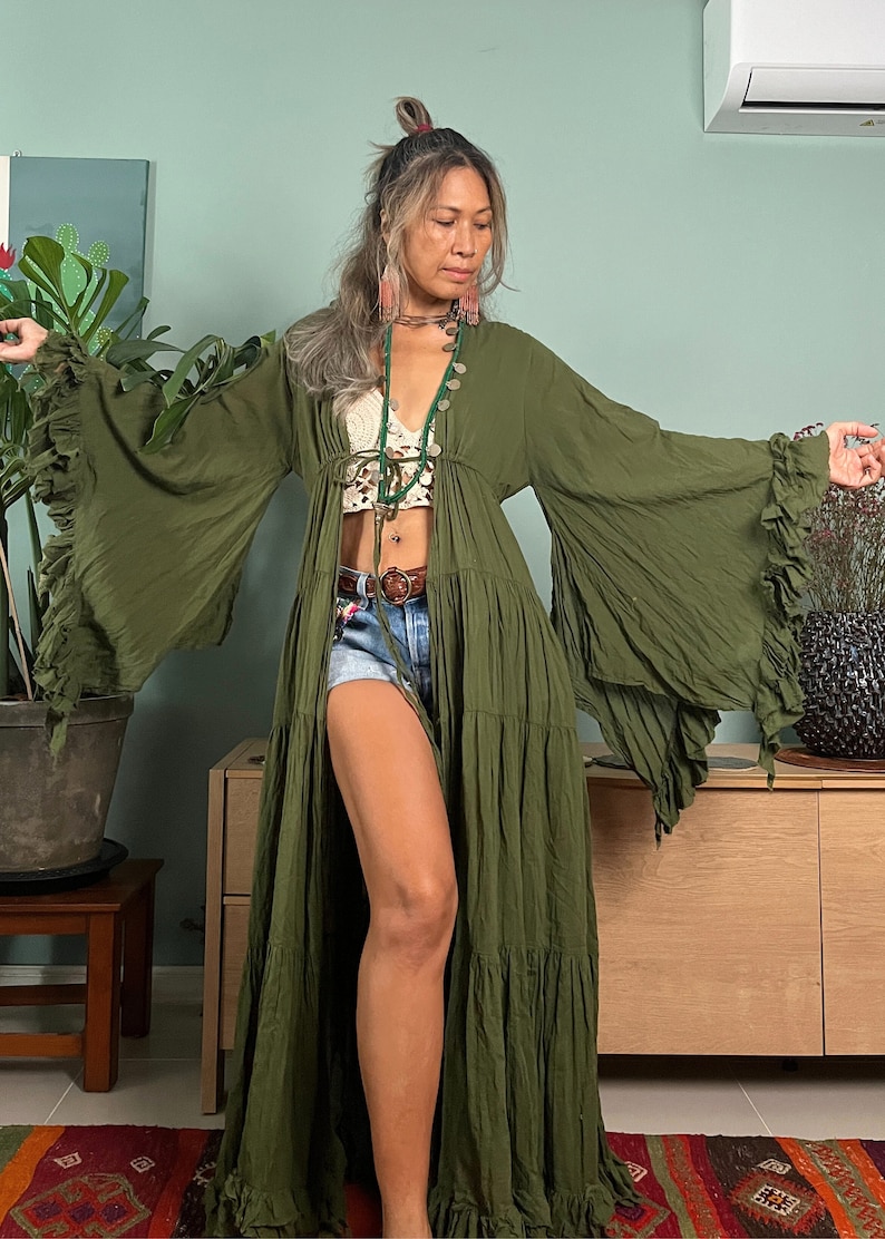 May include: Olive green maxi dress with long, ruffled sleeves. The dress is open in the front and has a tiered skirt. The model is wearing a white crochet top and denim shorts. A brown belt and necklace complete the look. The dress is a bohemian style.
