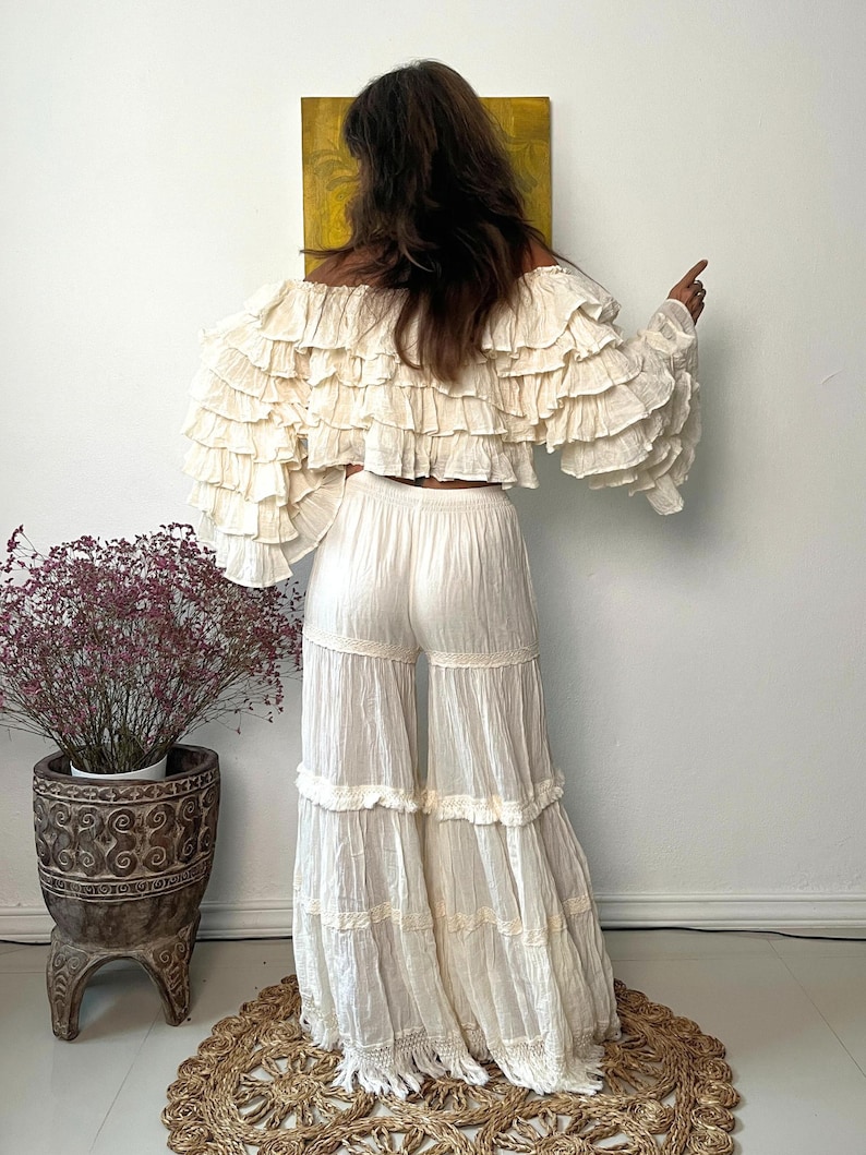 Pode incluir: Conjunto bo&ecirc;mio de duas pe&ccedil;as em branco sujo. A parte de cima &eacute; um top cropped com ombros de fora e babados em camadas. As cal&ccedil;as s&atilde;o de perna larga com babados em camadas e acabamento em renda. O conjunto &eacute; feito de um tecido leve. Um vaso decorativo com uma planta est&aacute; ao fundo.
