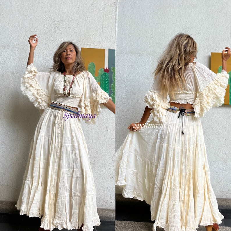May include: A white cotton peasant top with ruffled sleeves and a matching white cotton skirt. The top has a gathered bodice and a tie at the back. The skirt is long and flowing with multiple tiers. The model is wearing a beaded necklace and a belt with a colorful design.