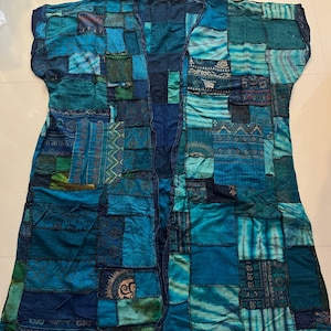 May include: A sleeveless open-front duster cardigan featuring a patchwork design in various shades of blue, teal, and green. The garment is constructed from rectangular fabric panels with diverse patterns, including geometric shapes and sun motifs. Text reads Style BU2.