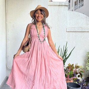 May include: A woman wearing a pink maxi dress with a straw hat and a silver and green necklace. She is standing on a tiled patio with a potted plant and a cup of coffee.