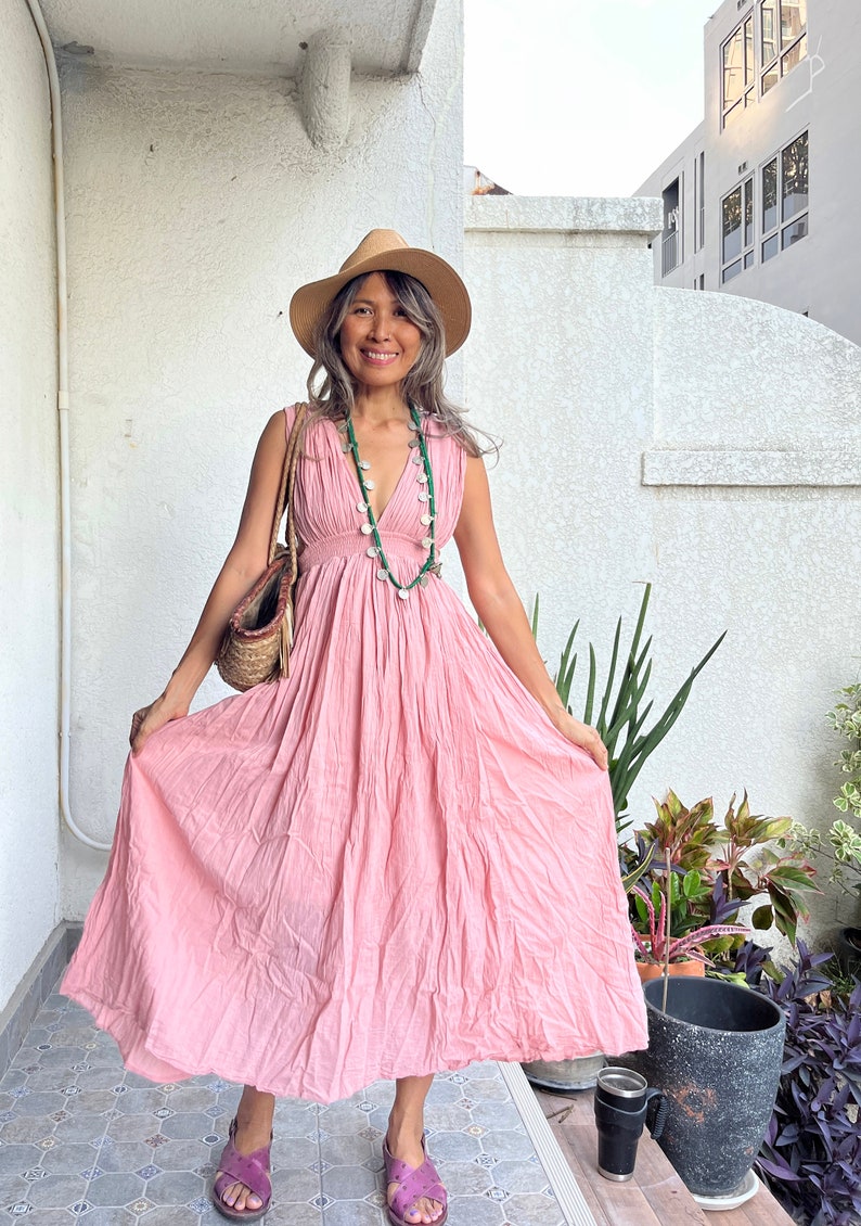 May include: A woman wearing a pink maxi dress with a straw hat and a silver and green necklace. She is standing on a tiled patio with a potted plant and a cup of coffee.