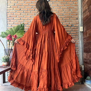 Op de afbeelding: Een oranje maxi-jurk met lange, wijde mouwen en ruches. De jurk heeft een getailleerde taille en een rok met ruches, wat een bohemian stijl cre&euml;ert. Gemaakt van een lichte stof, ideaal voor warm weer.