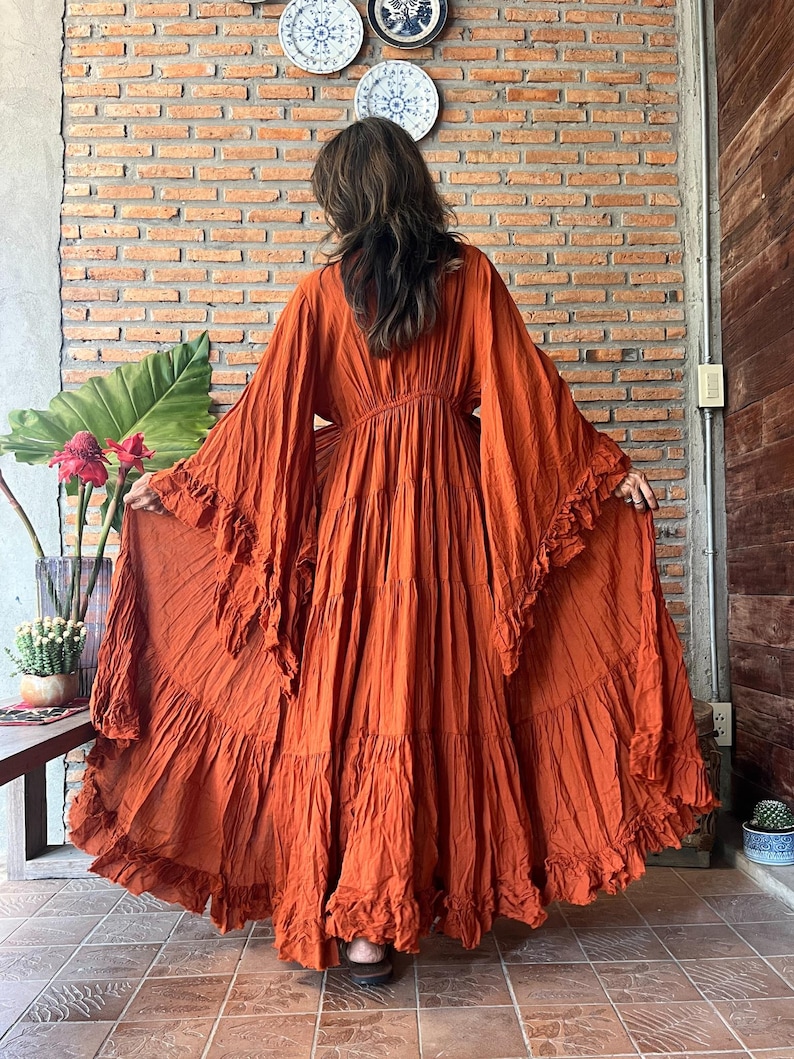 Op de afbeelding: Een oranje maxi-jurk met lange, wijde mouwen en ruches. De jurk heeft een getailleerde taille en een rok met ruches, wat een bohemian stijl cre&euml;ert. Gemaakt van een lichte stof, ideaal voor warm weer.