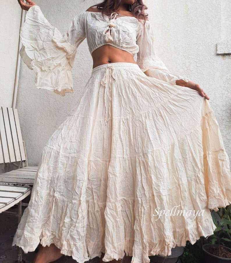 May include: A white off-the-shoulder crop top with long bell sleeves and a tie at the neckline, paired with a white tiered maxi skirt with a drawstring waist. The skirt has a ruffled hem and the text "Spellmaya" is visible on the bottom right corner of the skirt.