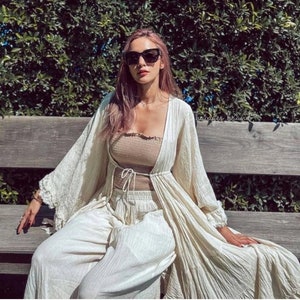 Op de afbeelding: Een vrouw draagt een beige tube top, een bijpassende broek en een lange, cr&egrave;mekleurige open cardigan. Ze draagt ook een zwarte zonnebril en zit op een houten bank voor een groene, bladerrijke achtergrond.